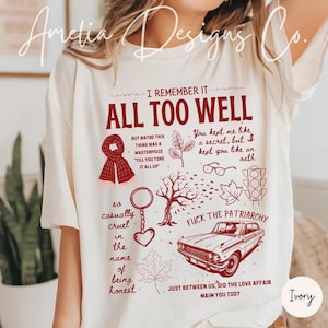 May include: Ivory t-shirt featuring a burgundy graphic design with text and illustrations. The design includes a scarf, glasses, a car, and maple leaves. The text reads "I Remember It All Too Well."
