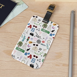 May include: White luggage tag with a colorful pattern of various icons, including a ghost, a snake, a guitar, sunglasses, and the text "Cornelia St" repeated multiple times.