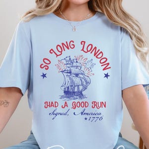 May include: Baby blue t-shirt with a nautical design featuring a ship, fireworks, and the text "So Long London, Had a Good Run, Signed, America *1776". The shirt also has the text "Amelia Designs Co."