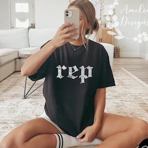 Może przedstawiać: Czarny t-shirt z nadrukiem słowa "rep" w białym kolorze w stylu gotyckim. Koszulkę nosi osoba siedząca na dywanie.