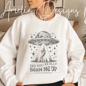 May include: White sweatshirt with a gray graphic of a UFO beaming a person up. The text "Did you really beam me up in a cloud of sparking dust" is printed below the graphic.
