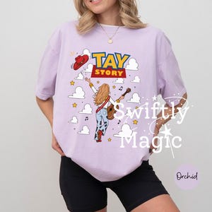 May include: A light purple t-shirt featuring a graphic of a person with long blonde hair wearing a red cowboy hat and cow-print pants, holding a guitar. The text TAY STORY is printed above, surrounded by white clouds, yellow stars, and musical notes.