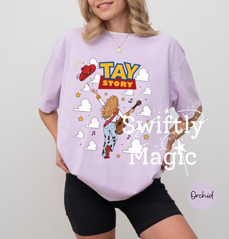 May include: A light purple t-shirt featuring a graphic of a person with long blonde hair wearing a red cowboy hat and cow-print pants, holding a guitar. The text TAY STORY is printed above, surrounded by white clouds, yellow stars, and musical notes.