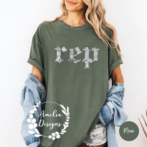 以下が含まれることがあります： 前面に白い文字で「REP」とプリントされた緑色のTシャツ。文字は新聞の印刷パターンで構成されています。シャツはデニムのショートパンツとデニムジャケットを着た人が着用しています。