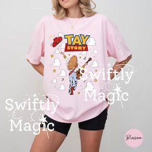 May include: A light pink t-shirt featuring a graphic print with the text TAY STORY. The design shows a person with long blonde hair wearing a white shirt, blue jeans with cow print, and red boots, holding a guitar, with a red cowboy hat above.