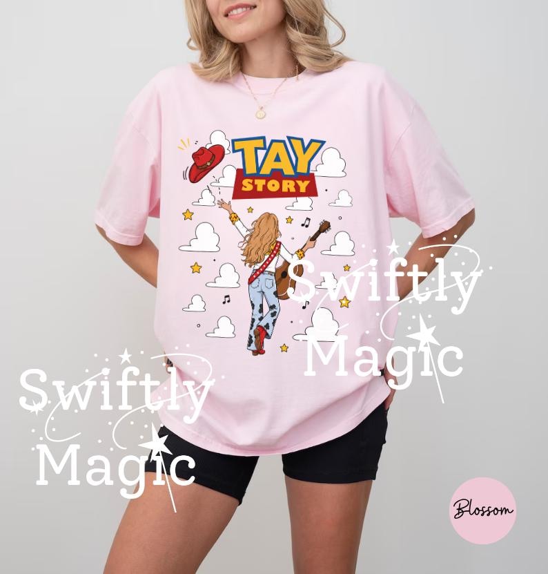 May include: A light pink t-shirt featuring a graphic print with the text TAY STORY. The design shows a person with long blonde hair wearing a white shirt, blue jeans with cow print, and red boots, holding a guitar, with a red cowboy hat above.