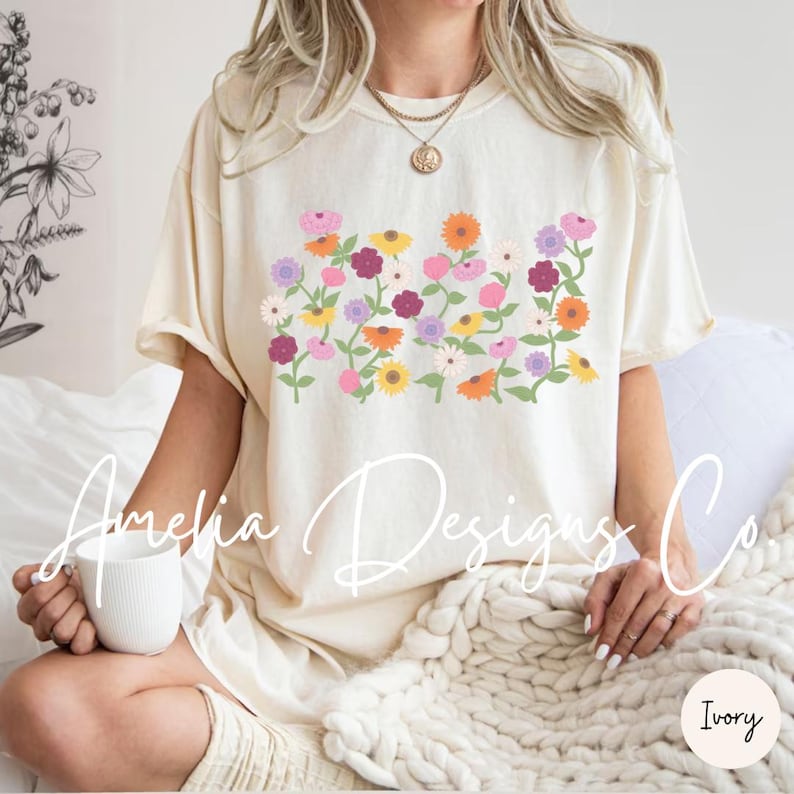 May include: A cream colored t-shirt with a floral design featuring pink, purple, yellow, orange, and green flowers. The text "Amelia Designs Co." is visible at the bottom of the shirt.