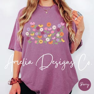 May include: A purple t-shirt with a floral design featuring a variety of colorful flowers. The text "Amelia Designs Co." is printed in white on the front of the shirt.