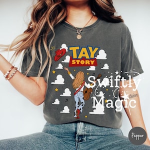 May include: A charcoal gray short-sleeve t-shirt featuring a graphic print with the text TAY STORY. The design includes a red cowboy hat, white clouds, yellow stars, and an illustration of a person with long hair holding a guitar.
