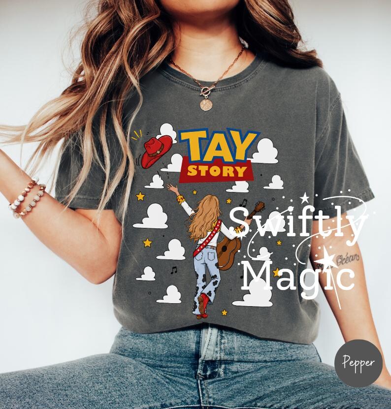 May include: A charcoal gray short-sleeve t-shirt featuring a graphic print with the text TAY STORY. The design includes a red cowboy hat, white clouds, yellow stars, and an illustration of a person with long hair holding a guitar.