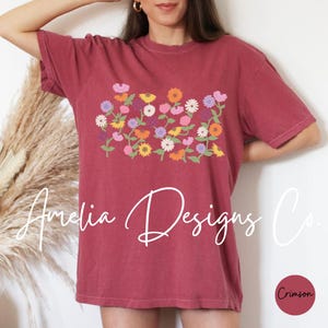 May include: A maroon t-shirt with a floral design featuring a variety of colorful flowers. The text "Amelia Designs Co." is printed in white on the bottom of the shirt.