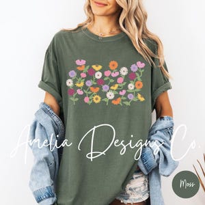 May include: A green t-shirt with a floral print featuring a variety of colorful flowers. The flowers are arranged in a horizontal row across the front of the shirt. The shirt has a crew neck and short sleeves.