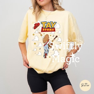May include: A pale yellow short-sleeve t-shirt featuring a graphic print with the text TAY STORY. The design includes a person with long hair holding a guitar, a red cowboy hat, and white clouds against a background of small yellow stars.