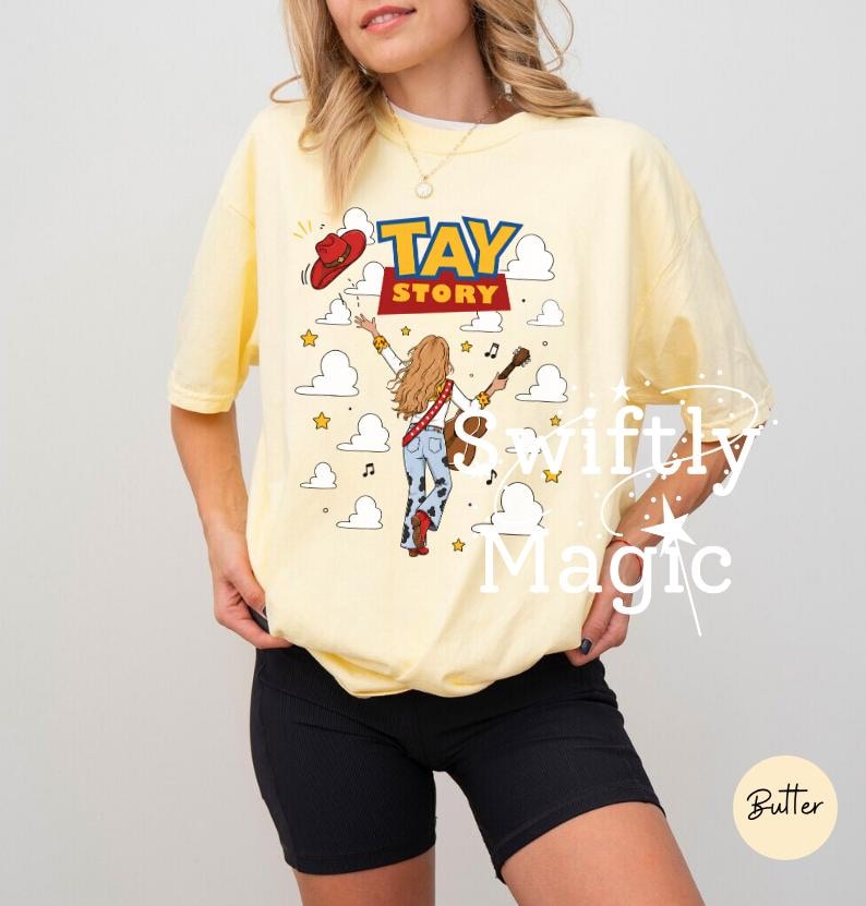 May include: A pale yellow short-sleeve t-shirt featuring a graphic print with the text TAY STORY. The design includes a person with long hair holding a guitar, a red cowboy hat, and white clouds against a background of small yellow stars.