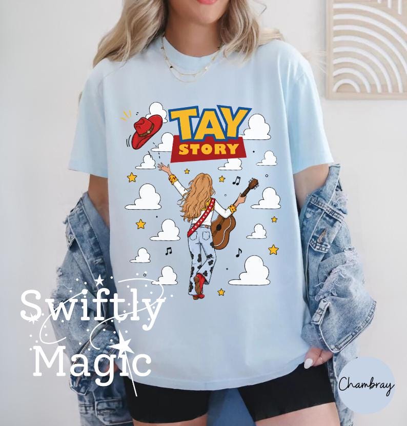 May include: A light blue t-shirt featuring a graphic of a person with long blonde hair playing an acoustic guitar. The design includes the text TAY STORY, a red cowboy hat, white clouds, yellow stars, and musical notes on a light blue background.