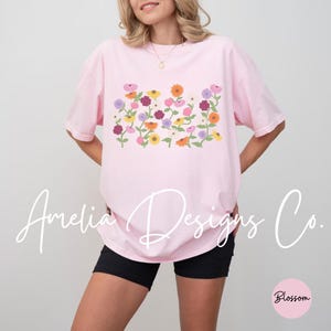 May include: A pink t-shirt with a floral design featuring a variety of colorful flowers. The shirt is loose-fitting and has a crew neck.