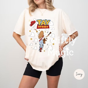 May include: An ivory t-shirt featuring a graphic print with the text TAY STORY in blue and red letters. The design includes a blonde figure with a guitar, a red cowboy hat, and cloud motifs. The figure wears blue jeans with cow print patterns.