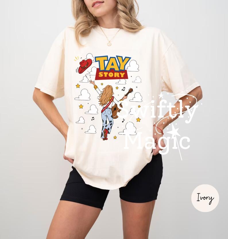 May include: An ivory t-shirt featuring a graphic print with the text TAY STORY in blue and red letters. The design includes a blonde figure with a guitar, a red cowboy hat, and cloud motifs. The figure wears blue jeans with cow print patterns.