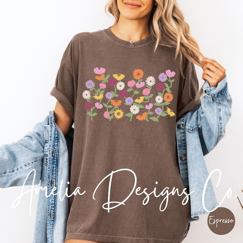 May include: A brown t-shirt with a floral design featuring a variety of colorful flowers. The text "Amelia Designs Co." is written in white cursive on the bottom left of the shirt. The text "Espresso" is written in white on a brown circle on the bottom right of the shirt.