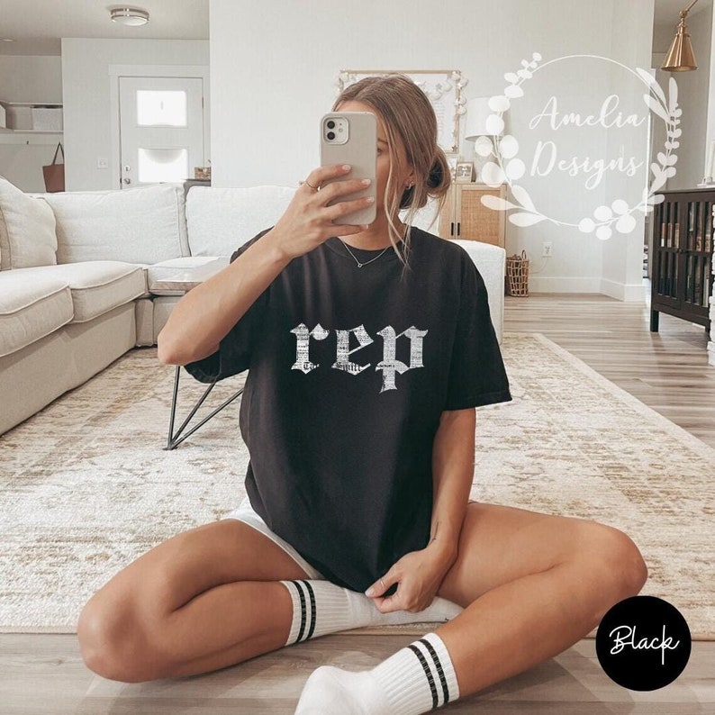 以下が含まれることがあります： ゴシック体で「rep」と白で書かれた黒いTシャツ。ラグに座っている人がモデルをしています。