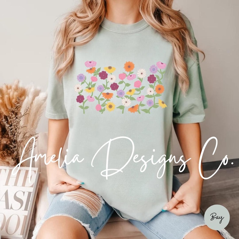 May include: A light blue t-shirt with a floral design featuring pink, purple, orange, and yellow flowers. The flowers are arranged in a horizontal row across the front of the shirt.