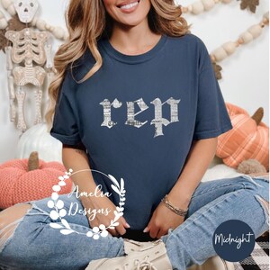 以下が含まれることがあります： 紺色のTシャツに、白で「rep」と古英語の文字で書かれた、擦れたようなグラフィックがプリントされています。このTシャツは、青いジーンズを履いた人が着用しています。