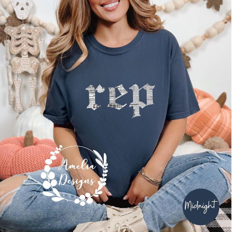以下が含まれることがあります： 紺色のTシャツに、白で「rep」と古英語の文字で書かれた、擦れたようなグラフィックがプリントされています。このTシャツは、青いジーンズを履いた人が着用しています。
