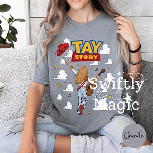 May include: A grey short-sleeve t-shirt featuring a graphic of a person with long hair playing an acoustic guitar, wearing cow-print pants and red boots. The design includes a red cowboy hat, white clouds, yellow stars, and the text TAY STORY.