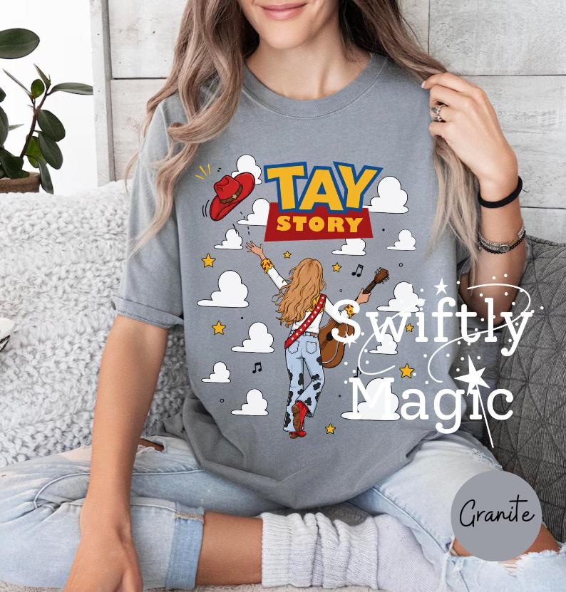 May include: A grey short-sleeve t-shirt featuring a graphic of a person with long hair playing an acoustic guitar, wearing cow-print pants and red boots. The design includes a red cowboy hat, white clouds, yellow stars, and the text TAY STORY.