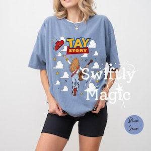 May include: A dusty blue short-sleeve t-shirt featuring a graphic print with the text TAY STORY. The design includes a person with long blonde hair playing a guitar, a red cowboy hat, yellow stars, musical notes, and white clouds.