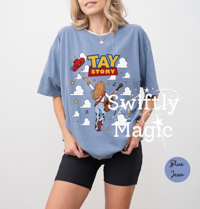 May include: A dusty blue short-sleeve t-shirt featuring a graphic print with the text TAY STORY. The design includes a person with long blonde hair playing a guitar, a red cowboy hat, yellow stars, musical notes, and white clouds.