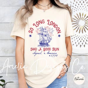 May include: Cream-colored t-shirt with a nautical design. The graphic features a blue and white ship, fireworks, and text that reads "So Long London, Had a Good Run, Signed, America, 1776."
