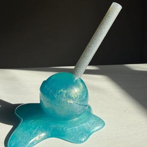 May include: A teal-colored resin lollipop sculpture with a glittery silver stick. The lollipop is round and sits on a matching teal puddle-shaped base. The sculpture is on a white surface with a dark background.