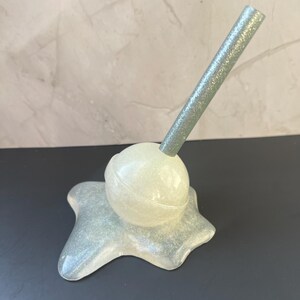 May include: A pearlescent, glittery, star-shaped object with a spherical top and a glittery, angled rod. The object is a light, iridescent color and sits on a dark surface.