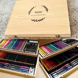 May include: A wooden art supply box with a light wood finish. The lid is engraved with the name "Katie" and the phrase "Let your creativity flow." Inside are multiple trays filled with coloured pencils, pens, and a pencil sharpener.
