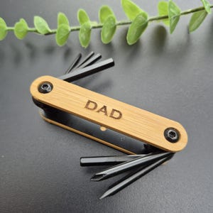 May include: A multi-tool with a bamboo handle engraved with the word "DAD". The tool features various black metal attachments, including Allen keys and screwdrivers. The tool is set against a dark background with a sprig of green leaves.