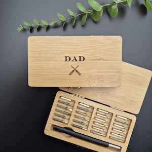 May include: A bamboo toolbox engraved with the word "DAD" and crossed screwdrivers on the lid. Inside, an array of screwdriver bits and a black handle are visible. The box is open on a dark surface.