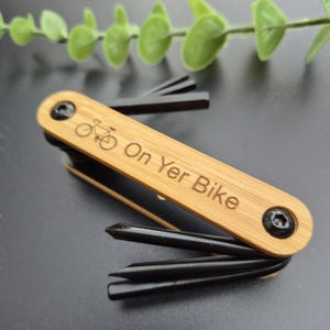 May include: A bamboo multi-tool with black tools, engraved with a bicycle and the words "On Yer Bike". This compact tool is designed for bicycle maintenance and repair, ideal for cyclists.