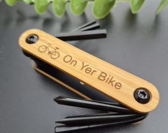 May include: A multi-tool with a bamboo handle and black tools. The handle is engraved with a bicycle and the words "On Yer Bike". The tool is designed for bicycle maintenance and repair.