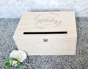 May include: A light-coloured wooden box with a slot on top, possibly for cards or gifts. The box is engraved with the names "Mr & Mrs Holliday" and the date "15 08 2023" within a floral wreath. A white flower with green leaves is in the foreground.