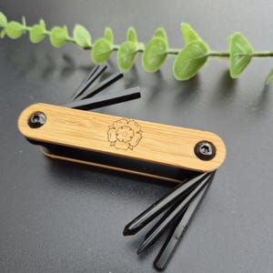 May include: A multi-tool with a bamboo handle and black tools. The handle has a laser-etched floral design. The tool is designed for various uses and is shown on a dark surface with a green plant in the background.