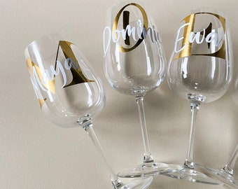 May include: Four clear glass wine glasses with gold and white lettering. The glasses are personalized with the names "Maya", "Dominic", "Ewan", and "Sarah".