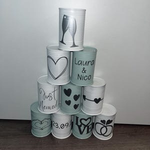 Könnte beinhalten: Ein Stapel aus acht weiß und grün lackierten Blechdosen mit verschiedenen Designs zum Thema Hochzeit. Die oberste Dose hat ein Paar Champagnergläser, die nächste Dose hat ein Herz, die nächste Dose steht "Laura & Nico", die nächste Dose steht "Just Married", die nächste Dose hat vier Herzen, die nächste Dose hat einen Pfeil und ein Herz, die nächste Dose steht "23.09.2", die nächste Dose steht "Love", und die unterste Dose hat zwei Ringe.