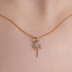 May include: A gold-toned necklace featuring a delicate chain and a small, slender palm tree pendant. The palm tree charm is encrusted with tiny, clear, sparkling stones that catch the light, suspended from a simple, thin, polished gold-colored metal chain.