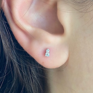 May include: A close-up of an ear with a small, delicate stud earring. The earring features two small, clear gemstones set close together. The skin tone is fair, and the earring is positioned on the earlobe.