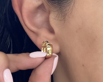 May include: A small, polished gold hoop earring with a thick, rounded profile. The earring features a hinged closure mechanism and a smooth, reflective surface that catches the light, creating a bright, metallic appearance on the earlobe.