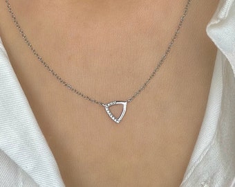 May include: A delicate silver necklace featuring a small, open triangular pendant. The pendant is embellished with small, sparkling stones. The necklace is worn against a white shirt, highlighting the jewelry's design and color.
