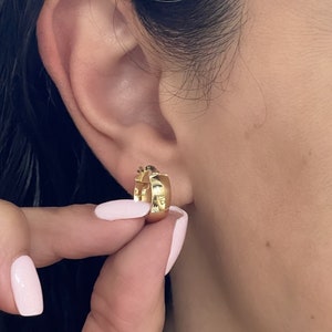 May include: A small, polished gold hoop earring with a thick, rounded profile. The earring features a hinged closure mechanism and a smooth, reflective surface that catches the light, creating a bright, metallic appearance on the earlobe.