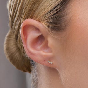 May include: Close-up of a person's ear with three small, sparkling stud earrings. The earrings are set in a row. The person has blonde hair pulled back in a bun. The background is a soft, neutral color.