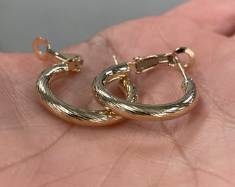 May include: A pair of gold-colored hoop earrings with a textured, ridged design. The earrings are round and have a clasp closure. The earrings are shown on a light-colored background.